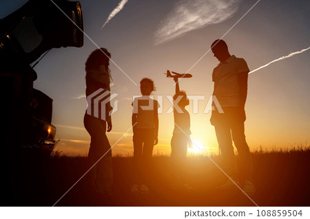 Black silhouettes of family members fly kite near car Black silhouettes of family members fly kite near car 108859504