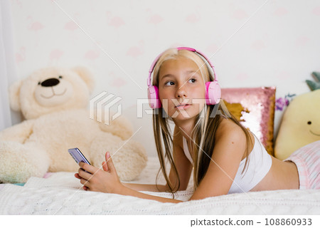 Beautiful teenage girl is relaxing in bed using a touchscreen smartphone, listening to music with a wireless headset, surfing the Internet in the bedroom, relaxing comfortably, smiling, enjoying a Beautiful teenage girl is relaxing in bed using a touchscreen smartphone, listening to music with a wireless headset, surfing the Internet in the bedroom, relaxing comfortably, smiling, enjoying a 108860933