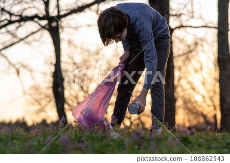 Teenage boy collects garbage in big pink bag in nature, on meadow on autumn evening at sunset. Environmental problems. 108862543