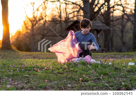 Children's volunteer activity. Teenage boy collects garbage in large pink bag in nature on autumn evening at sunset. 108862545