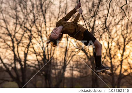 Happy teenage boy flies high up on homemade swing in nature, in park, in village against background of autumn trees. 108862546