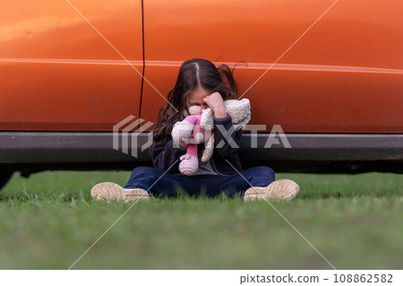 Little lonely girl is crying, hiding her face in toy, sitting on grass with her back pressed against car, upset. 108862582