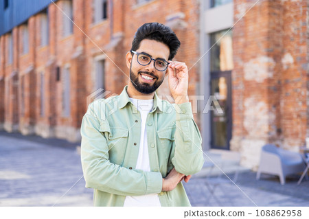 Portrait of young man in casual clothes joyful businessman smiling and looking at camera with arms crossed, Indian man outside office building. 108862958