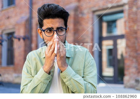 A man sneezes while sitting on a bench outside an office building, a young businessman has a runny nose and allergies, sick. A man sneezes while sitting on a bench outside an office building, a young businessman has a runny nose and allergies, sick. 108862960
