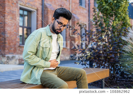A sick man is sitting on a bench outside an office building near a park, he has severe pain in his stomach, an Indian man is upset and holds his stomach with his hands. A sick man is sitting on a bench outside an office building near a park, he has severe pain in his stomach, an Indian man is upset and holds his stomach with his hands. 108862962