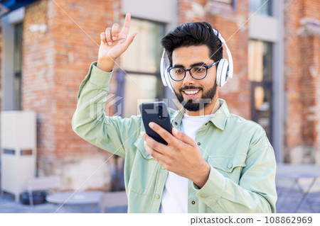 A young man in headphones is walking around the city, joyful, smiling, listening to music online, using an application on the phone. A young man in headphones is walking around the city, joyful, smiling, listening to music online, using an application on the phone. 108862969