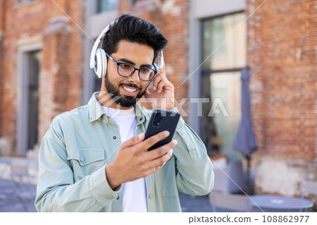 A young man in headphones is walking around the city, joyful, smiling, listening to music online, using an application on the phone. 108862977