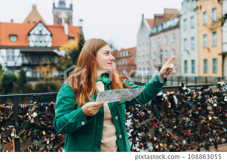 Young pretty 30s woman with backpack holding paper map and pointing to object on copy space, rear view against the Millers' Guild house and the Radunia Canal in Gdansk, 108863055