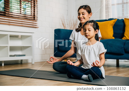 In a cozy home setting a young mother and her charming daughter practice family yoga in lotus position emphasizing mindfulness and meditation with smiles reflecting togetherness and happiness. In a cozy home setting a young mother and her charming daughter practice family yoga in lotus position emphasizing mindfulness and meditation with smiles reflecting togetherness and happiness. 108863132