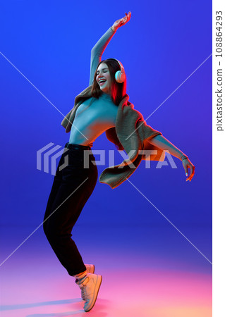 Overjoyed, happy girl, student dancing while listening music in headphones against blue studio background in neon light, filter. Overjoyed, happy girl, student dancing while listening music in headphones against blue studio background in neon light, filter. 108864293