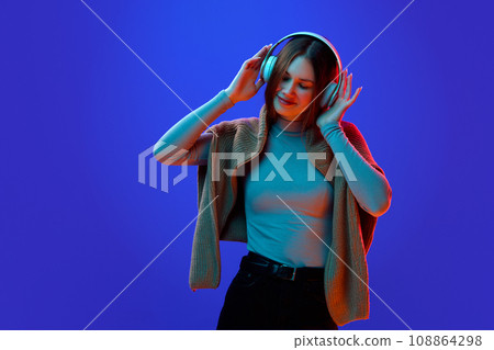 Portrait of young attractive girl, student listening music in headphones against blue studio background in neon light, filter. 108864298