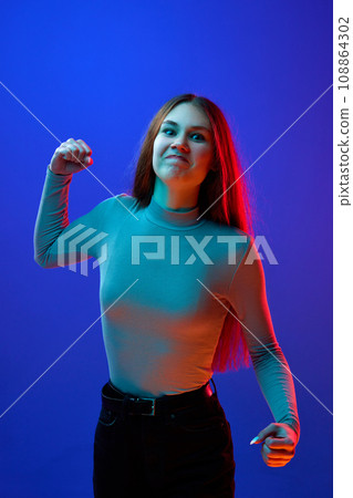 Young attractive woman standing looking at camera arguing and showing, clenching fist against blue background in neon light filter. Young attractive woman standing looking at camera arguing and showing, clenching fist against blue background in neon light filter. 108864302