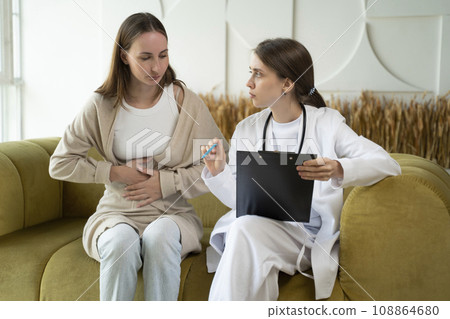 A patient with abdominal pain undergoes a medical examination by a doctor about an upset stomach. A young woman is talking to a doctor and holding her stomach 108864680