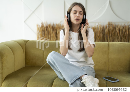 Carefree brunette listens to music and sings with headphones while sitting on the sofa in the living room. Happy young lady with closed eyes enjoys music, home interior 108864682