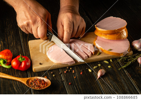 Slicing parboiled sausage with a knife in the hands of a chef for preparing a delicious lunch on the kitchen table. Fast food concept Slicing parboiled sausage with a knife in the hands of a chef for preparing a delicious lunch on the kitchen table. Fast food concept 108866685