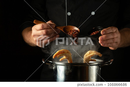 The cook adds aromatic seasoning to a pot of simmering chicken legs. Concept of preparing delicious soup or broth in the kitchen of a restaurant or hotel The cook adds aromatic seasoning to a pot of simmering chicken legs. Concept of preparing delicious soup or broth in the kitchen of a restaurant or hotel 108866686