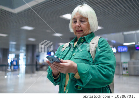 Travel, airport senior 60s woman in comfy casual outfit going vacation alone, holding a smartphone and booking cheap flights online Travel, airport senior 60s woman in comfy casual outfit going vacation alone, holding a smartphone and booking cheap flights online 108867015