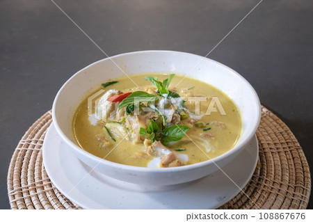 Green Curry with Chicken garnished on top with pepper and basil was served with white bowl put on wicker cream color placemat. Green Curry with Chicken garnished on top with pepper and basil was served with white bowl put on wicker cream color placemat. 108867676