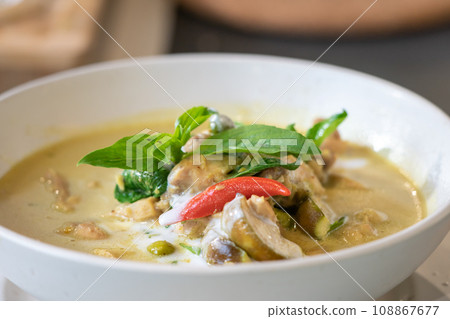 Close up to Green Curry with Chicken garnished on top with pepper and basil was served with white bowl put on wicker cream color placemat. 108867677
