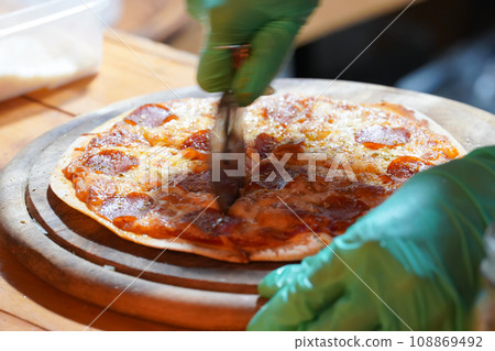 A person wearing green gloves is cutting thin-crust, small-size pizzas on a wooden tray with a pizza cutter. 108869492