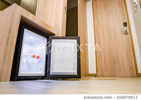 Small refrigerator under a wooden counter with two water bottles in a hotel room. This minimalist fridge comes in black and white color with LED and 3 shelves inside. 108870011