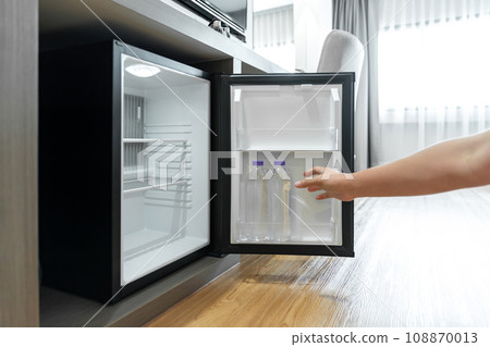 Man Hand opened mini small refrigerator and reached drinking plastic water bottle under the wood counter in the hotel resort bedroom. 108870013
