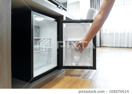Man Hand opened mini small refrigerator and reached drinking plastic water bottle under the wood counter in the hotel resort bedroom. 108870014