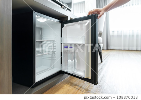 Man Hand opened mini small refrigerator and reached drinking plastic water bottle under the wood counter in the hotel resort bedroom. Man Hand opened mini small refrigerator and reached drinking plastic water bottle under the wood counter in the hotel resort bedroom. 108870018