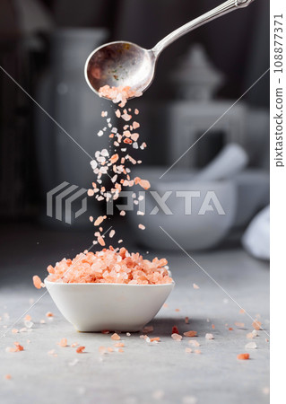 Pink salt in crystals is poured into a small white bowl. 108877371