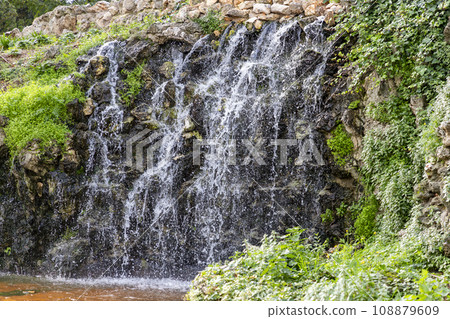 Waterfall. Waterfall in a park in Madrid. Artificial Mountain of Buen Retiro, called Mountain of the Cats. Dark stone background surrounded by green vegetation and plants and leaves. Autumn colors. 108879609