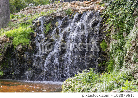 Waterfall. Waterfall in a park in Madrid. Artificial Mountain of Buen Retiro, called Mountain of the Cats. Dark stone background surrounded by green vegetation and plants and leaves. Autumn colors. 108879615