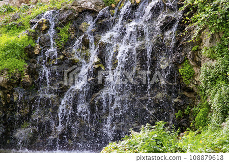 Waterfall. Waterfall in a park in Madrid. Artificial Mountain of Buen Retiro, called Mountain of the Cats. Dark stone background surrounded by green vegetation and plants and leaves. Autumn colors. 108879618