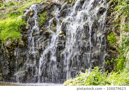Waterfall. Waterfall in a park in Madrid. Artificial Mountain of Buen Retiro, called Mountain of the Cats. Dark stone background surrounded by green vegetation and plants and leaves. Autumn colors. 108879623