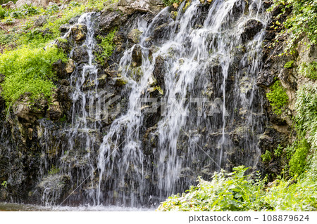 Waterfall. Waterfall in a park in Madrid. Artificial Mountain of Buen Retiro, called Mountain of the Cats. Dark stone background surrounded by green vegetation and plants and leaves. Autumn colors. Waterfall. Waterfall in a park in Madrid. Artificial Mountain of Buen Retiro, called Mountain of the Cats. Dark stone background surrounded by green vegetation and plants and leaves. Autumn colors. 108879624