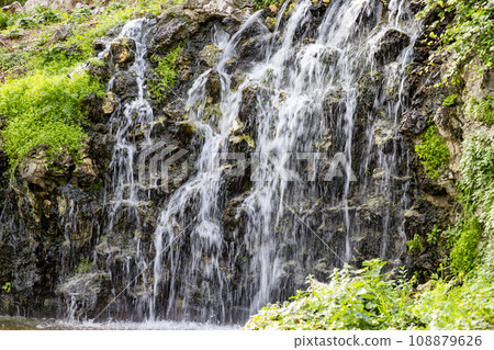 Waterfall. Waterfall in a park in Madrid. Artificial Mountain of Buen Retiro, called Mountain of the Cats. Dark stone background surrounded by green vegetation and plants and leaves. Autumn colors. Waterfall. Waterfall in a park in Madrid. Artificial Mountain of Buen Retiro, called Mountain of the Cats. Dark stone background surrounded by green vegetation and plants and leaves. Autumn colors. 108879626