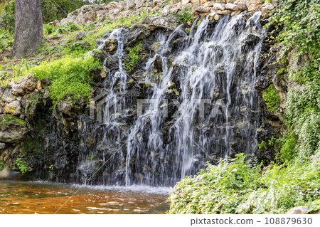Waterfall. Waterfall in a park in Madrid. Artificial Mountain of Buen Retiro, called Mountain of the Cats. Dark stone background surrounded by green vegetation and plants and leaves. Autumn colors. 108879630