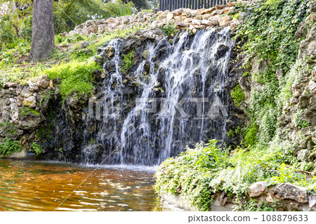 Waterfall. Waterfall in a park in Madrid. Artificial Mountain of Buen Retiro, called Mountain of the Cats. Dark stone background surrounded by green vegetation and plants and leaves. Autumn colors. 108879633