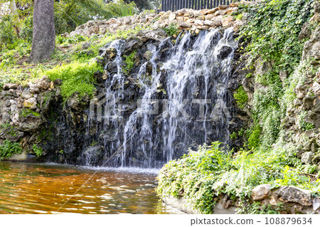 Waterfall. Waterfall in a park in Madrid. Artificial Mountain of Buen Retiro, called Mountain of the Cats. Dark stone background surrounded by green vegetation and plants and leaves. Autumn colors. 108879634
