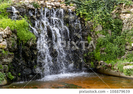 Waterfall. Waterfall in a park in Madrid. Artificial Mountain of Buen Retiro, called Mountain of the Cats. Dark stone background surrounded by green vegetation and plants and leaves. Autumn colors. 108879639