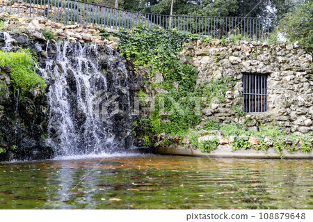 Waterfall. Waterfall in a park in Madrid. Artificial Mountain of Buen Retiro, called Mountain of the Cats. Dark stone background surrounded by green vegetation and plants and leaves. Autumn colors. 108879648