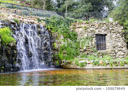Waterfall. Waterfall in a park in Madrid. Artificial Mountain of Buen Retiro, called Mountain of the Cats. Dark stone background surrounded by green vegetation and plants and leaves. Autumn colors. 108879649