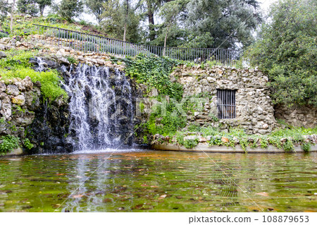 Waterfall. Waterfall in a park in Madrid. Artificial Mountain of Buen Retiro, called Mountain of the Cats. Dark stone background surrounded by green vegetation and plants and leaves. Autumn colors. 108879653