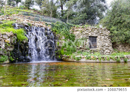 Waterfall. Waterfall in a park in Madrid. Artificial Mountain of Buen Retiro, called Mountain of the Cats. Dark stone background surrounded by green vegetation and plants and leaves. Autumn colors. Waterfall. Waterfall in a park in Madrid. Artificial Mountain of Buen Retiro, called Mountain of the Cats. Dark stone background surrounded by green vegetation and plants and leaves. Autumn colors. 108879655
