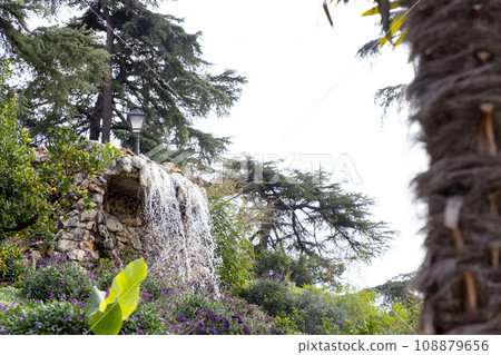 Waterfall. Waterfall in a park in Madrid. Artificial Mountain of Buen Retiro, called Mountain of the Cats. Dark stone background surrounded by green vegetation and plants and leaves. Autumn colors. Waterfall. Waterfall in a park in Madrid. Artificial Mountain of Buen Retiro, called Mountain of the Cats. Dark stone background surrounded by green vegetation and plants and leaves. Autumn colors. 108879656