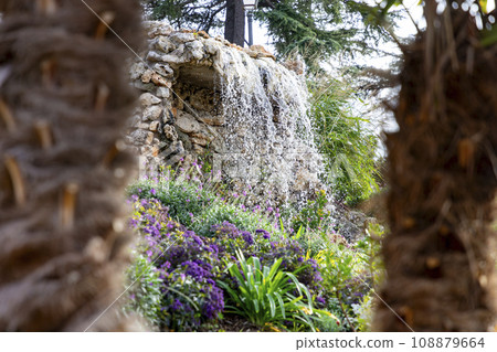 Waterfall. Waterfall in a park in Madrid. Artificial Mountain of Buen Retiro, called Mountain of the Cats. Dark stone background surrounded by green vegetation and plants and leaves. Autumn colors. Waterfall. Waterfall in a park in Madrid. Artificial Mountain of Buen Retiro, called Mountain of the Cats. Dark stone background surrounded by green vegetation and plants and leaves. Autumn colors. 108879664