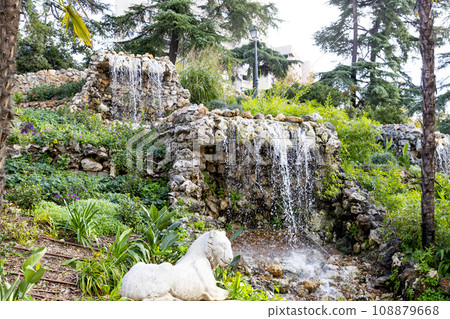 Waterfall. Waterfall in a park in Madrid. Artificial Mountain of Buen Retiro, called Mountain of the Cats. Dark stone background surrounded by green vegetation and plants and leaves. Autumn colors. 108879668