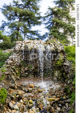 Waterfall. Waterfall in a park in Madrid. Artificial Mountain of Buen Retiro, called Mountain of the Cats. Dark stone background surrounded by green vegetation and plants and leaves. Autumn colors. 108879671