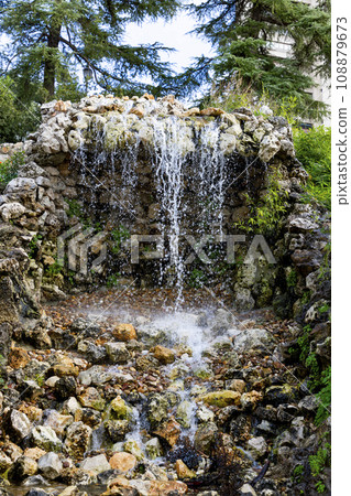 Waterfall. Waterfall in a park in Madrid. Artificial Mountain of Buen Retiro, called Mountain of the Cats. Dark stone background surrounded by green vegetation and plants and leaves. Autumn colors. 108879673