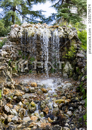 Waterfall. Waterfall in a park in Madrid. Artificial Mountain of Buen Retiro, called Mountain of the Cats. Dark stone background surrounded by green vegetation and plants and leaves. Autumn colors. 108879674