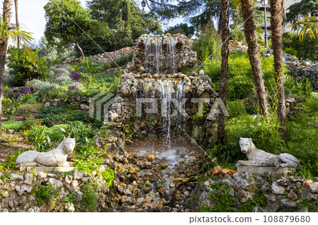 Waterfall. Waterfall in a park in Madrid. Artificial Mountain of Buen Retiro, called Mountain of the Cats. Dark stone background surrounded by green vegetation and plants and leaves. Autumn colors. 108879680
